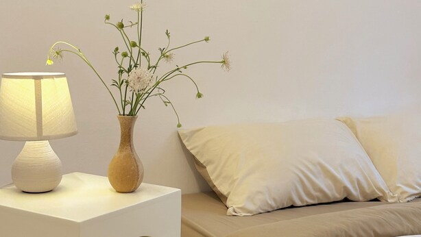 A bedroom with a bedside table, a lamp, a vase with flowers, and a bed with pillows. - TRIP HOME stay & studio (Ho Chi Minh City)