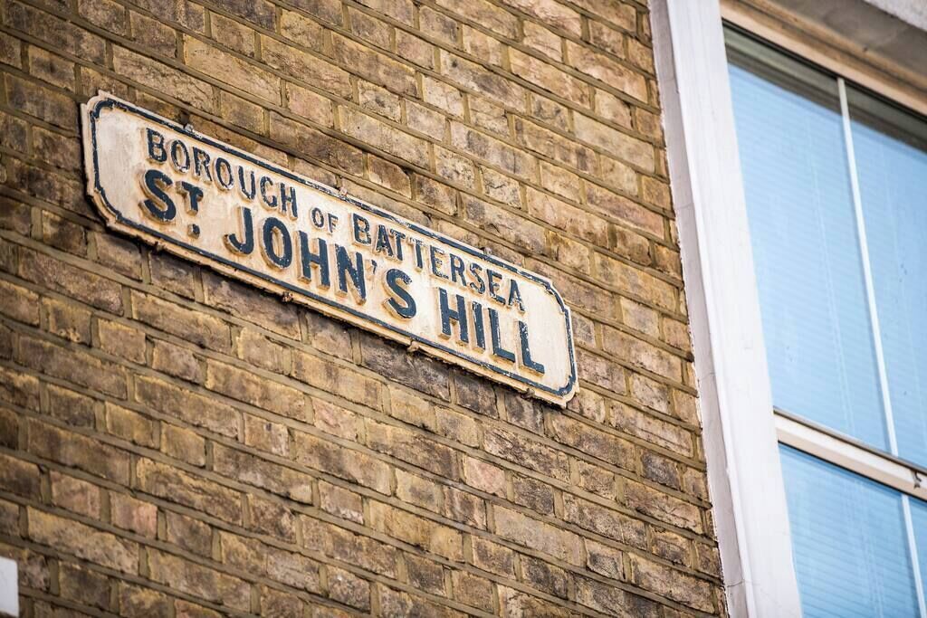 Een verweerd straatnaambord op een bakstenen muur, met de tekst 'Borough of Battersea St. John's Hill'.