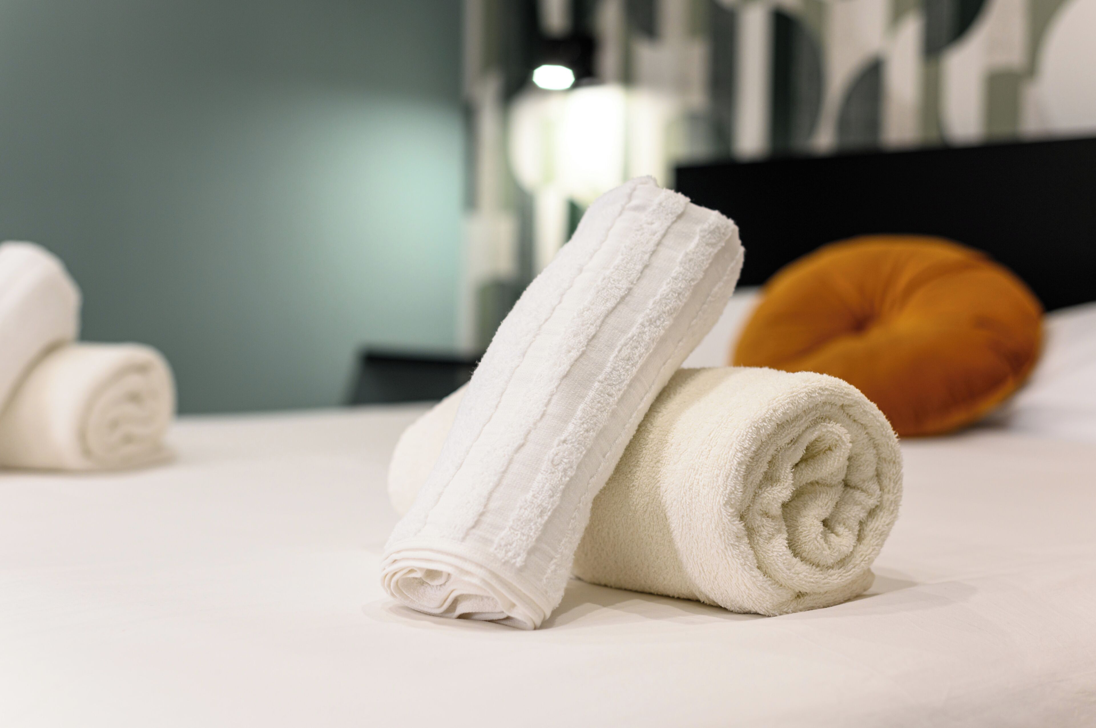 A neatly made bed with two rolled white towels and a single orange pillow.