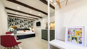 Une chambre moderne avec un lit, une table de chevet et un téléviseur fixé au mur. On y trouve également un coin repas avec une table et des chaises, ainsi qu’une cuisine équipée de placards et d’un plan de travail.