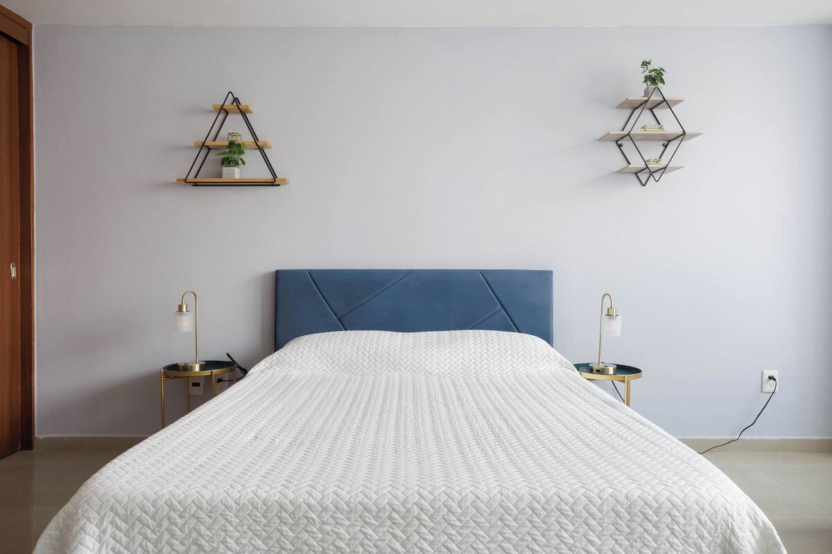 A bedroom with a bed, a blue headboard, bedside tables, and wall-mounted shelves with plants.