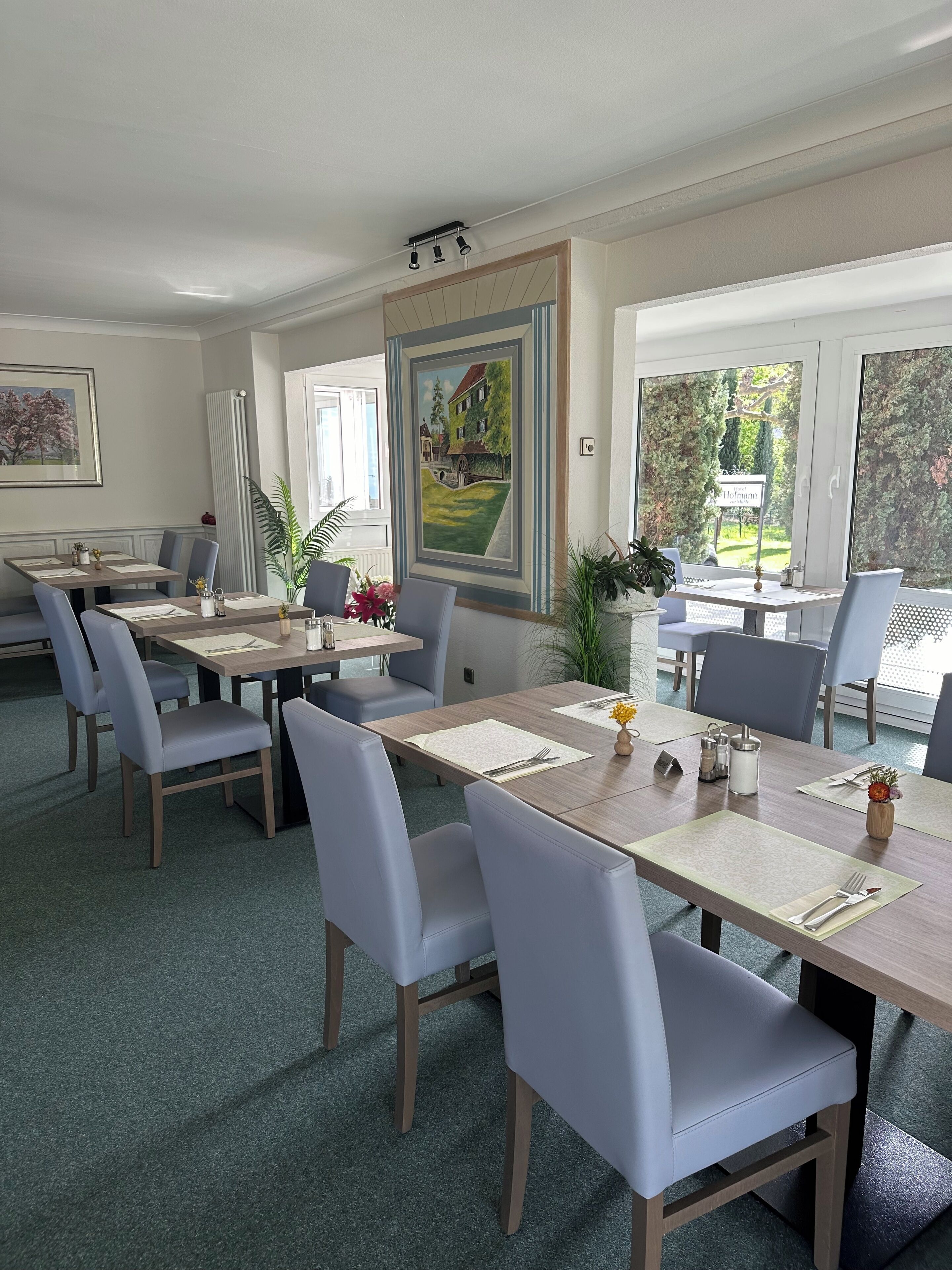 A dining area with multiple tables and chairs, framed artwork on the walls, and a view of greenery outside.