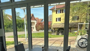 Un invernadero con una mesa y sillas, ventanas amplias y vistas a un edificio amarillo y una rueda hidráulica.