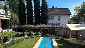 Una piscina rectangular con superficie de agua azul cristalina, rodeada por una terraza de piedra. Hay sillas de descanso y una pérgola cubierta de enredaderas con flores moradas.