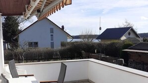 A rooftop terrace with a table and chairs, a covered area with a corrugated roof, and a view of neighboring houses and trees.