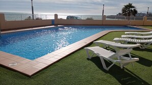 Una piscina con bordes azulejados, rodeada de césped artificial y sillas reclinables blancas.