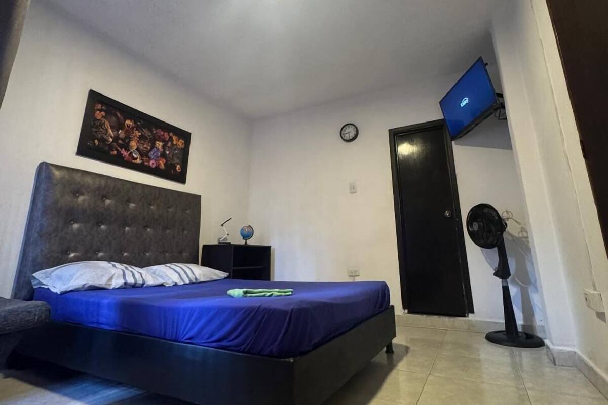 A bedroom with a bed, a nightstand, a clock, a wall-mounted TV, and a standing fan.