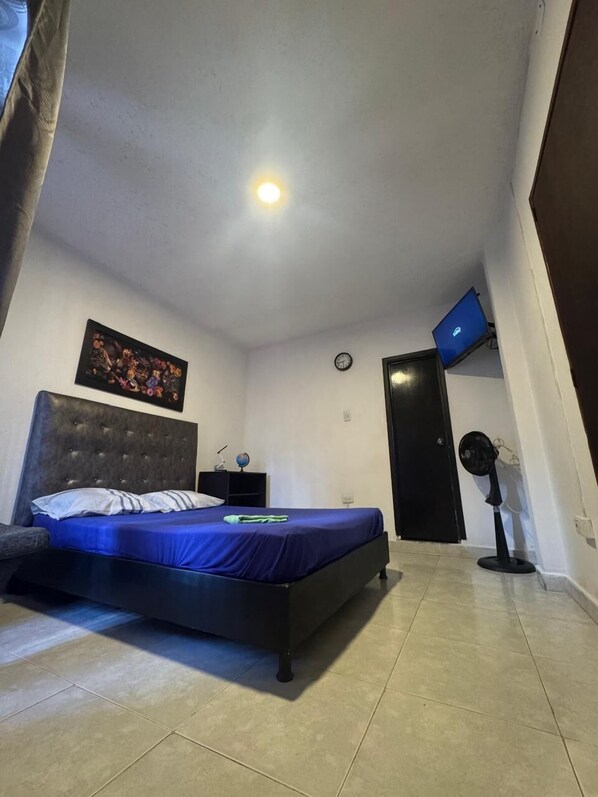 A bedroom with a bed, a nightstand, a clock, a wall-mounted TV, and a standing fan.