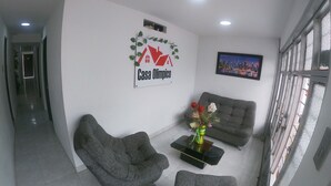 Ett modernt vardagsrum med en soffa, en fåtölj och ett soffbord med en vas med blommor. En målning som föreställer en stadsbild hänger på väggen, och en skylt visar texten "Casa Olimpico".
