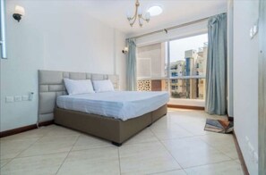 A bedroom with a bed, a chandelier, a window with curtains, and a view of buildings.
