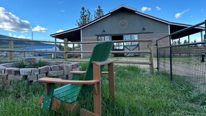 A rustic cabin with a large window and a wheel emblem, surrounded by a wooden fence and a grassy area with a chair. - Beautiful mountain views, Close to trails, Pet friendly Farm Awaits you! (Florence)