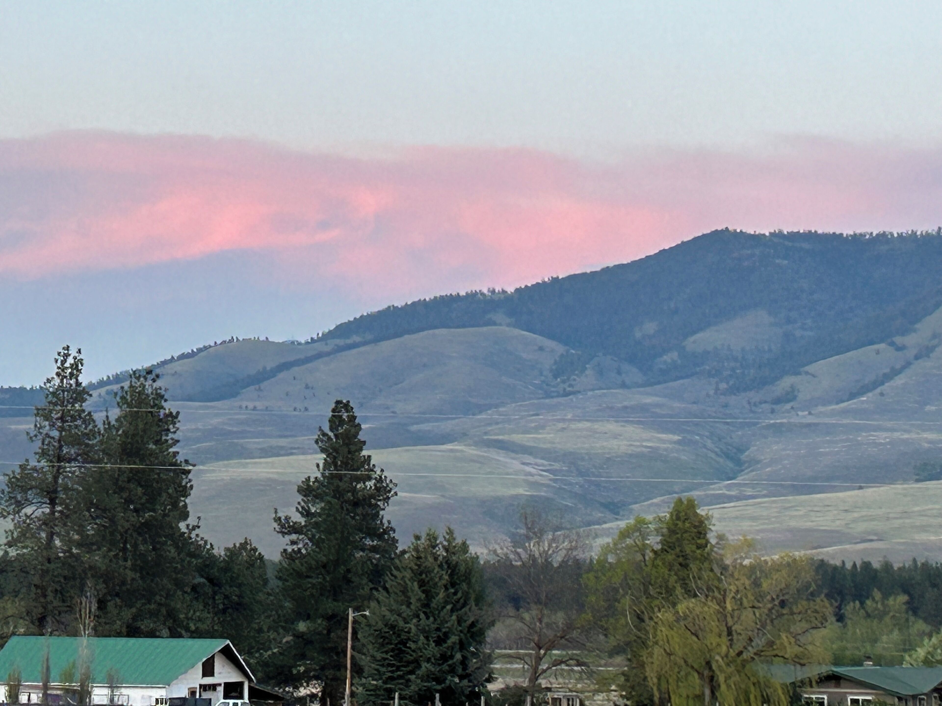 A rural landscape with rolling hills, a few houses with green roofs, and a clear sky with pink clouds.