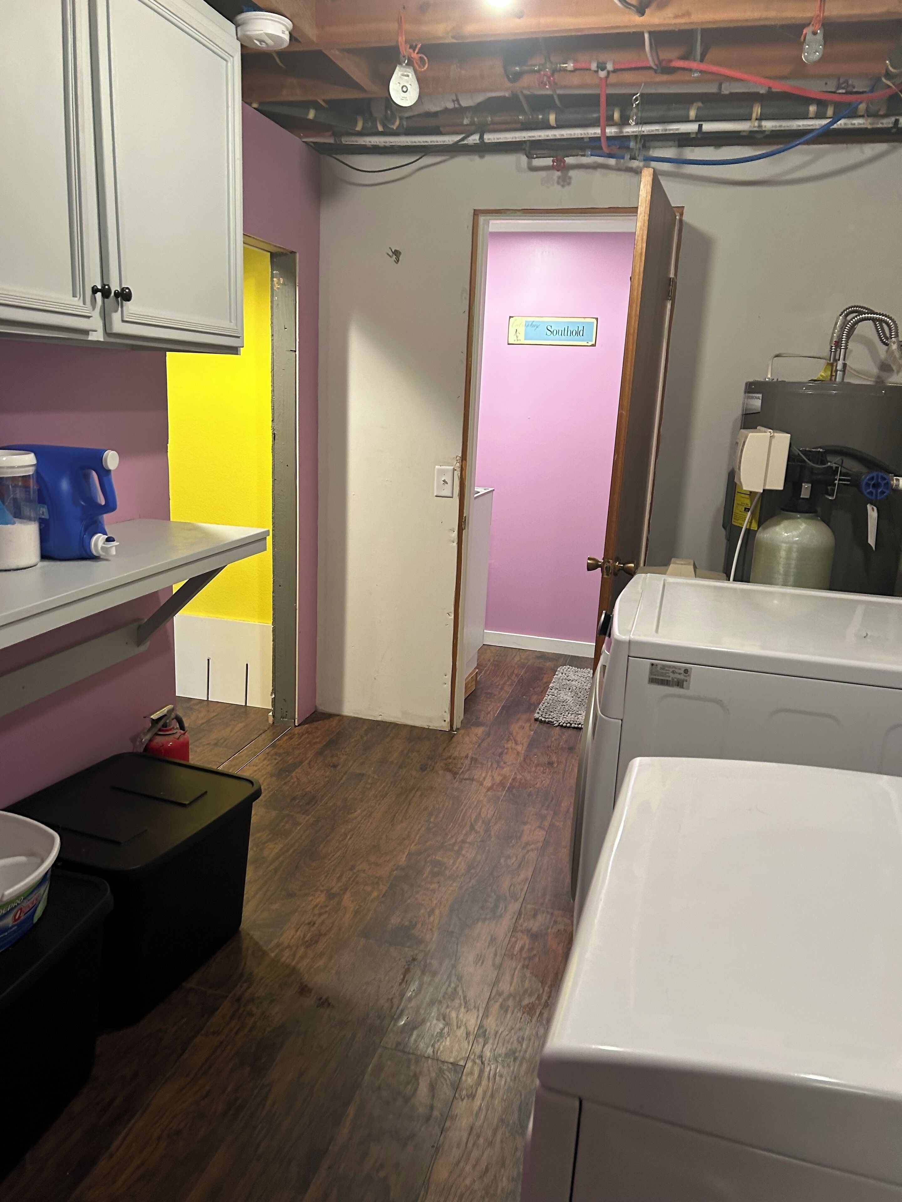 A laundry room with a washing machine, a utility sink, and a water heater. There is a door labeled "Southhold" and a shelf with a blue container.