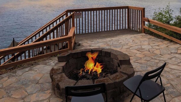 A stone deck with a fire pit and two chairs, overlooking a lake with a wooden railing and a view of the forested hills. - Beachfront House Getaway with Amazing Views&Access (Port Orchard)