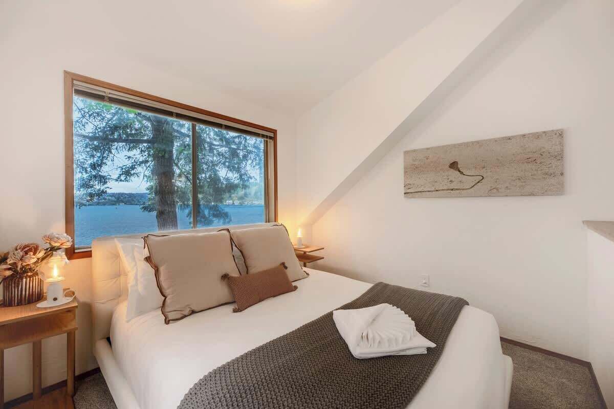 Une chambre avec une grande fenêtre, un lit avec des oreillers, une table de chevet avec une lampe et une œuvre d’art murale.