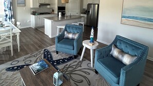 A modern living room with two blue armchairs, a wooden coffee table, and a dining area with a white table and chairs. A kitchen with white cabinets and stainless steel appliances is visible in the background. - Large, beautiful house! 1.5 miles to Bethany Beach! Resort style amenities!!! (Ocean View)