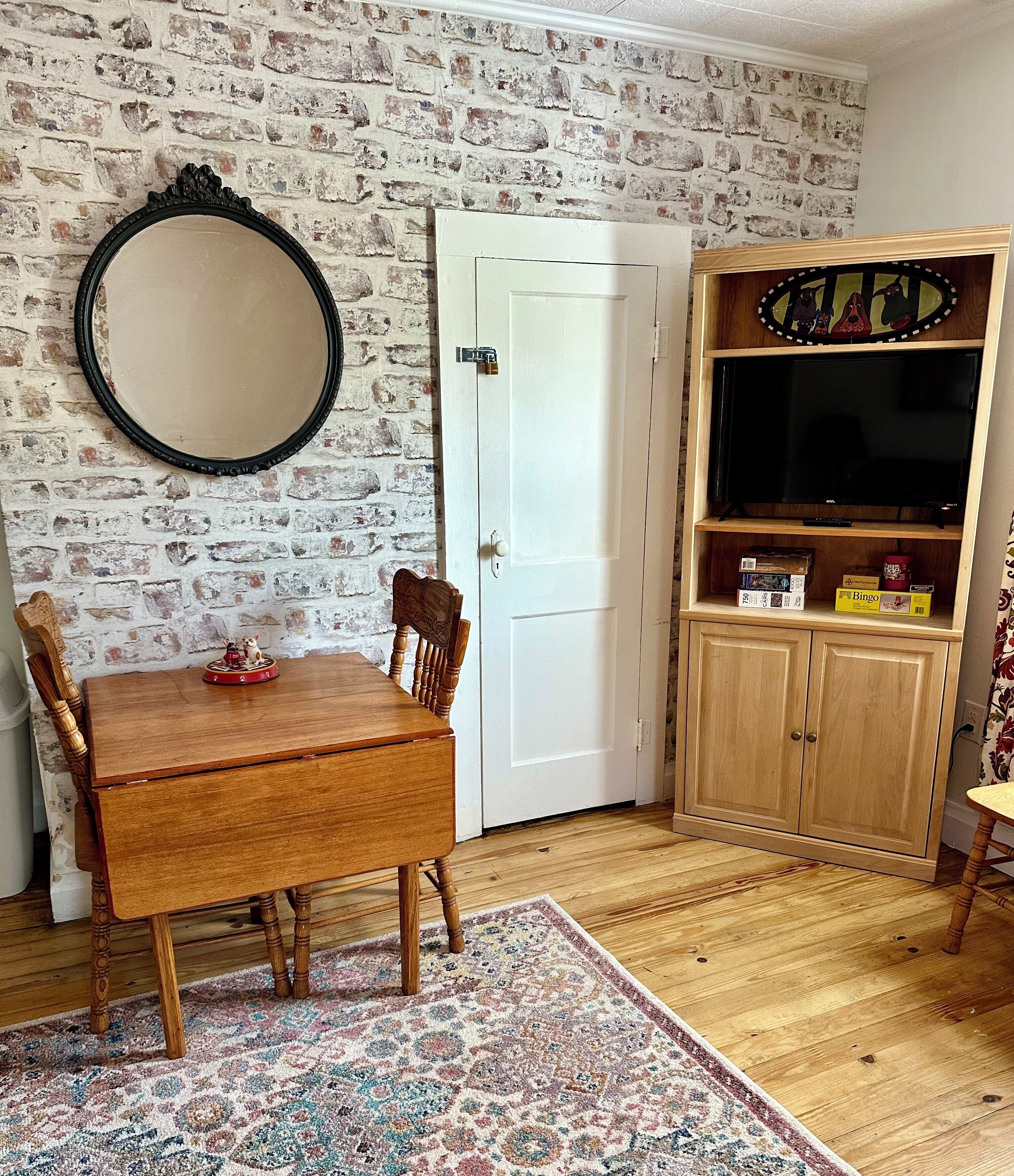 Una habitación con una mesa de comedor de madera, sillas, un tapete estampado, un espejo, una puerta blanca y un mueble de madera con un televisor.