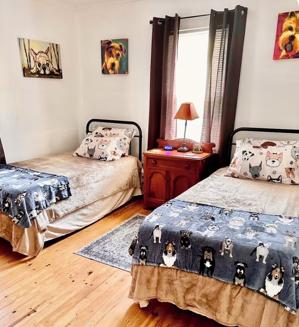 A bedroom with two single beds, each with a dog-themed duvet cover. There are two paintings on the wall, a nightstand with a lamp, and a window with curtains.