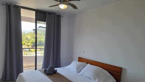 A bedroom with a bed, a wooden nightstand, a ceiling fan, and a view of greenery outside. - TAHITI OCEAN VIEW HOME (Papeete)