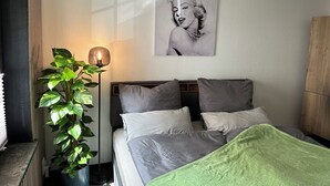 Un dormitorio con cama, almohadas, una manta verde, una planta en maceta, una lámpara de pie y un cartel en la pared.