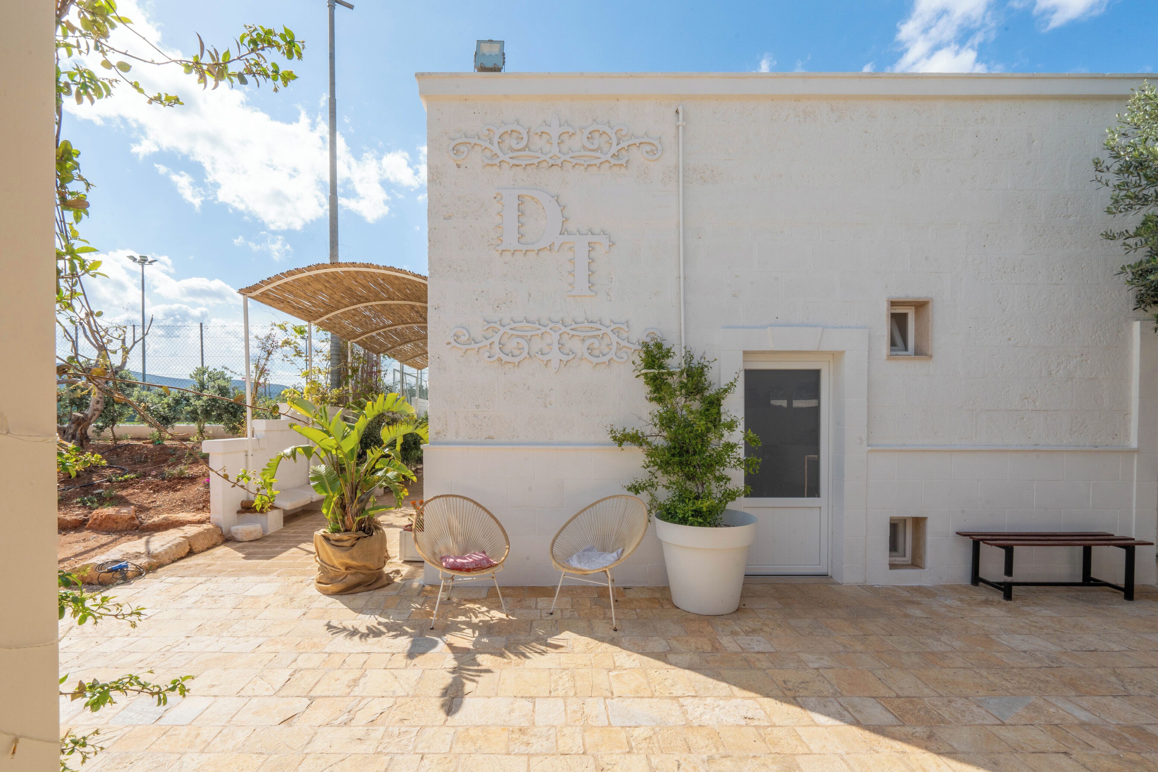 A courtyard with a white wall featuring a decorative monogram, two wicker chairs, a large potted plant, and a bench.