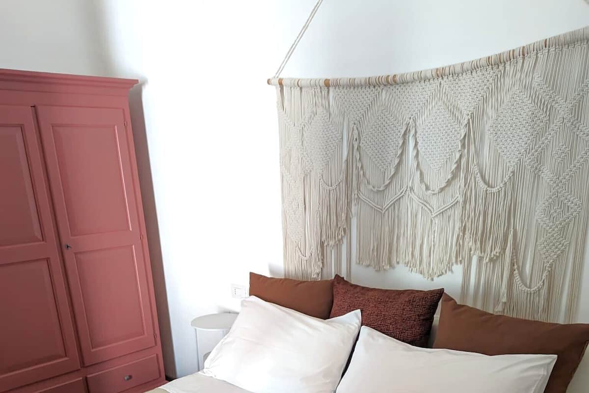 Una cama con tablero de cabecera de macramé, un armario rosa y una mesita de noche blanca.