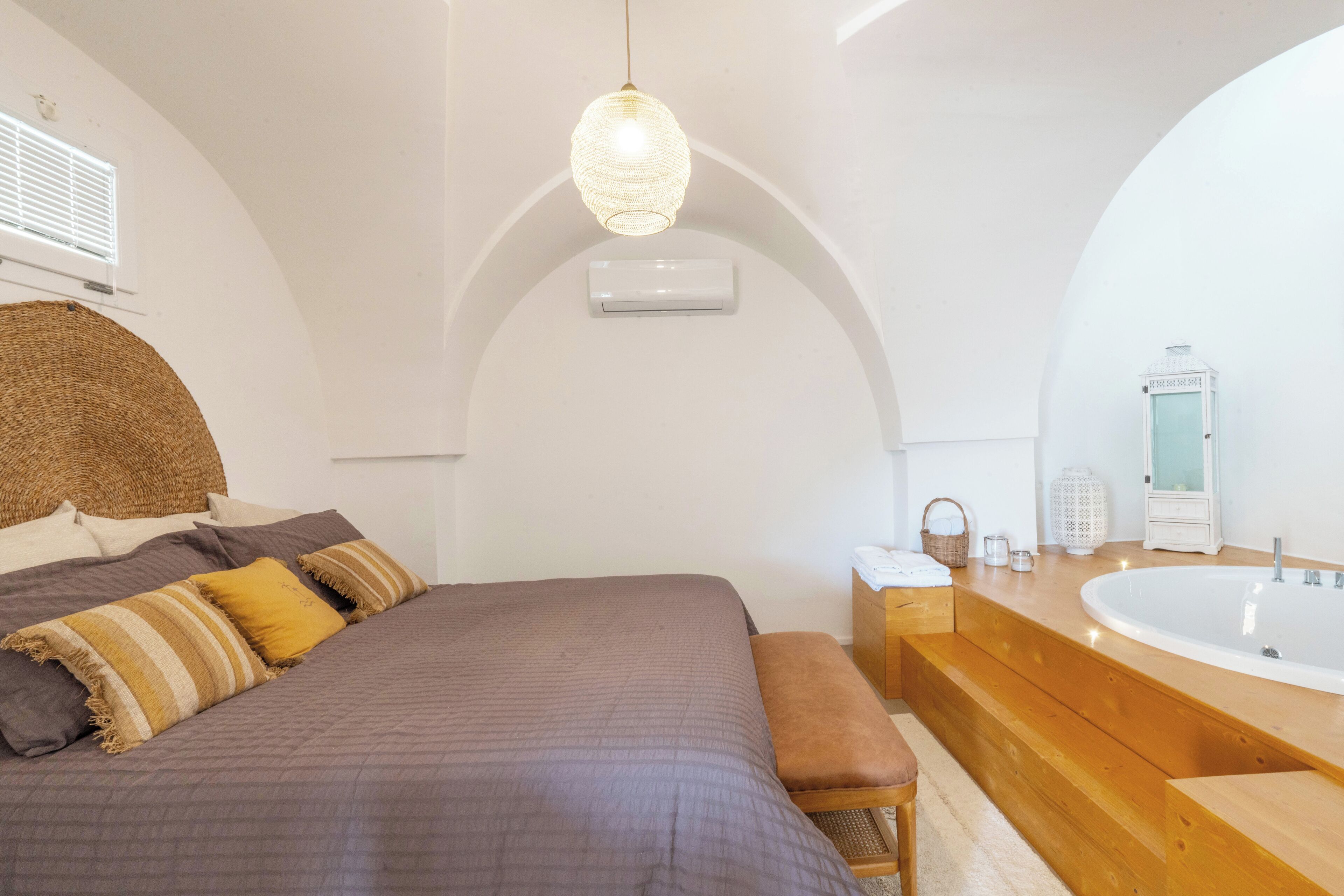 Ein Schlafzimmer mit einem großen Bett, einer hölzernen Bank, einer Pendelleuchte und einem Badezimmer mit Badewanne und Waschbecken.