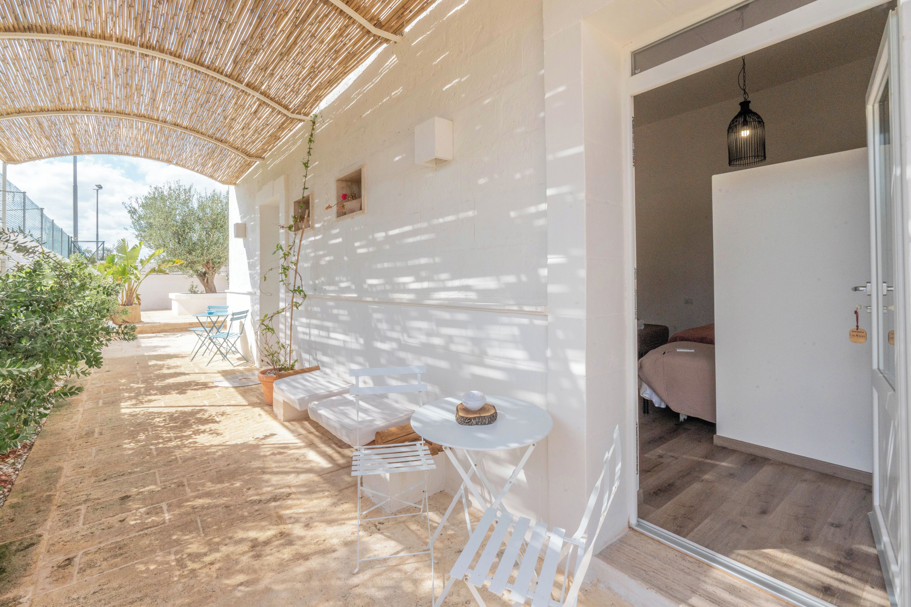 A covered outdoor area with a table and chairs, a door leading to a bedroom, and a view of the outside.
