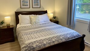 Un lit bien fait, recouvert d’une couvre-lit à motifs, deux œuvres d’art encadrées accrochées au mur, une lampe de chevet et une fenêtre avec des rideaux.