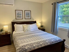 Una cama bien hecha con una colcha estampada, dos cuadros en la pared, una lámpara de noche y una ventana con cortinas.