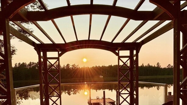 Une terrasse en bois surmontée d’une pergola, offrant une vue sur une étendue d’eau paisible au coucher du soleil.