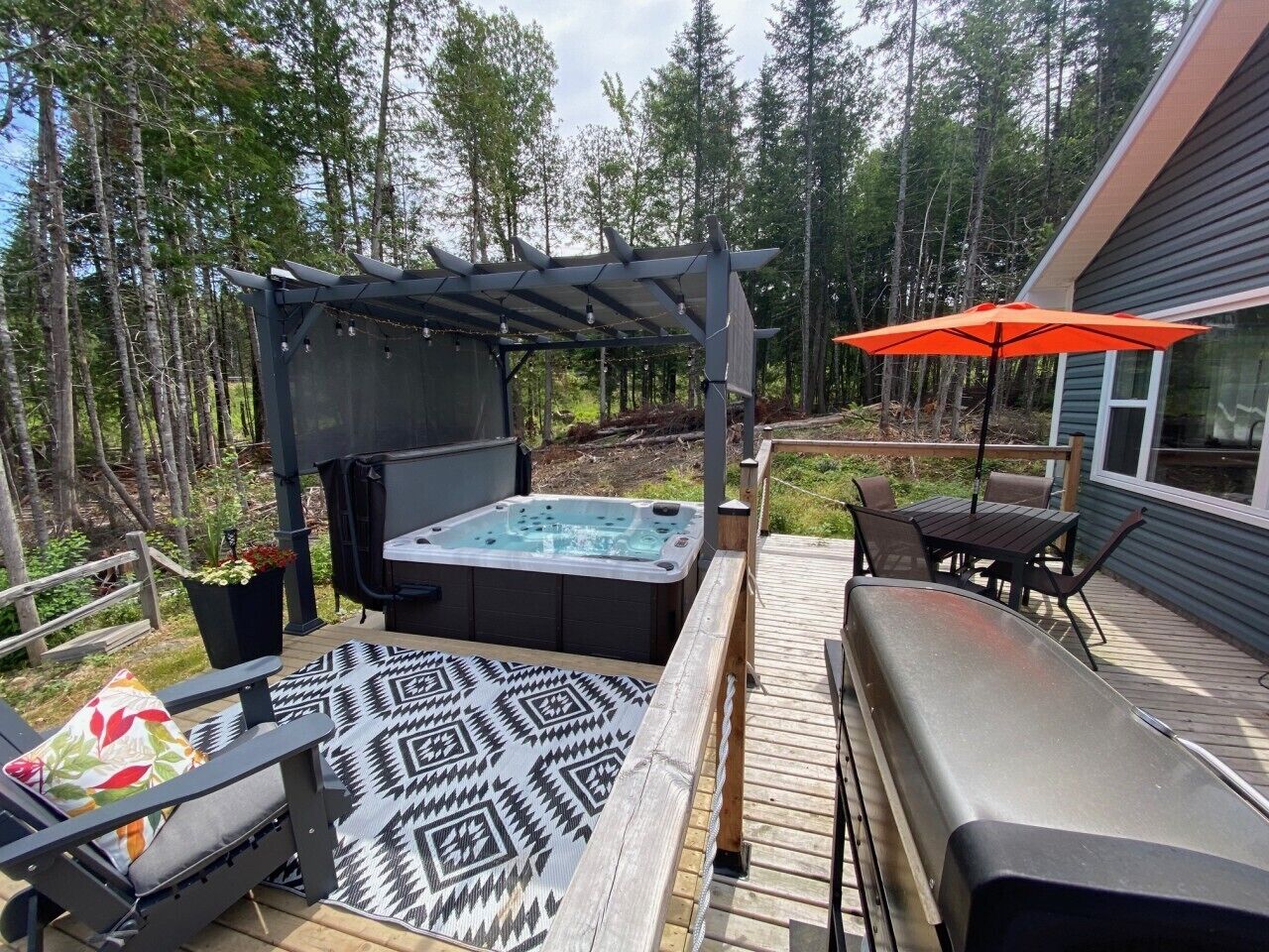 Et spabad på en træterrasse med et pergolaoverdækning, udendørs møbler og udsigt til et skovområde.