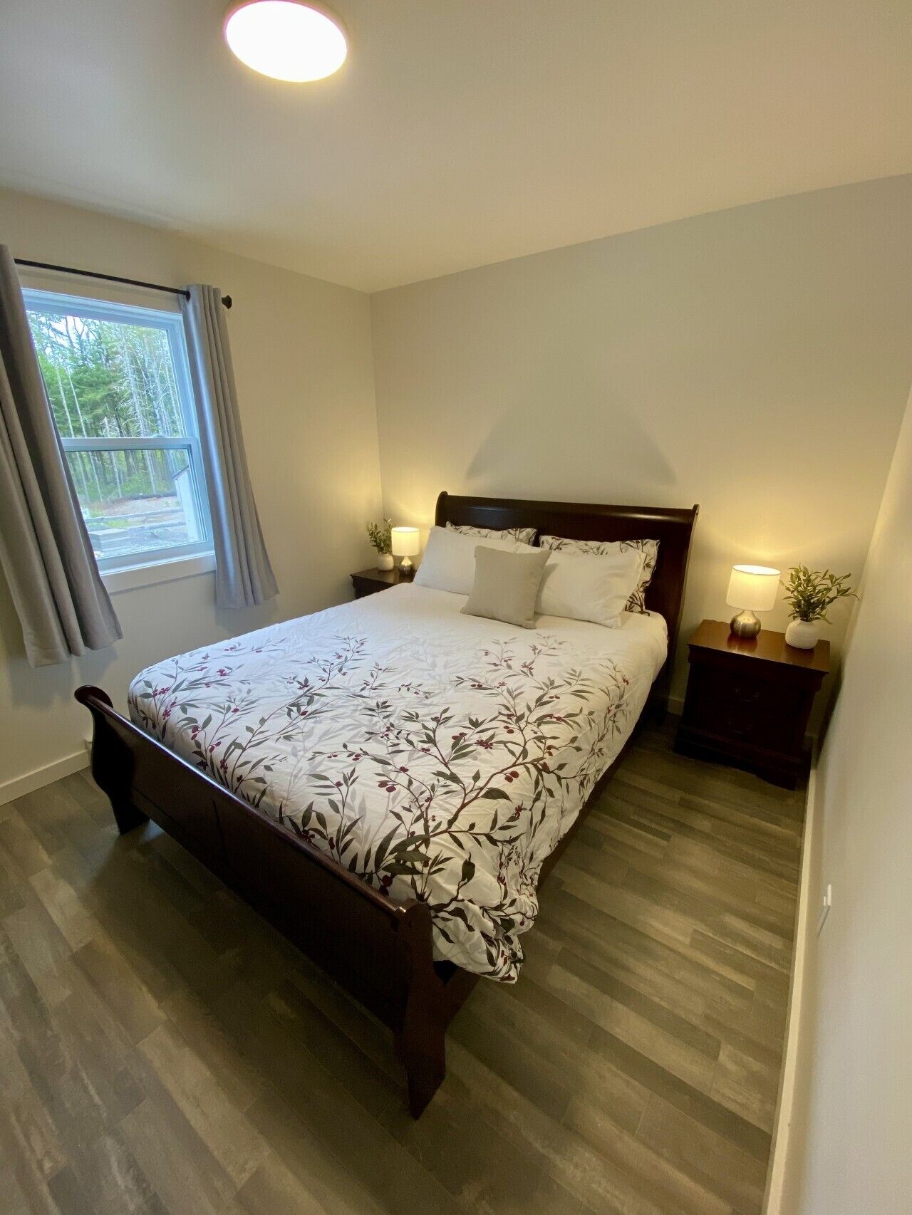 Une chambre à coucher avec un lit, deux tables de chevet, une fenêtre avec des rideaux et un luminaire au plafond.
