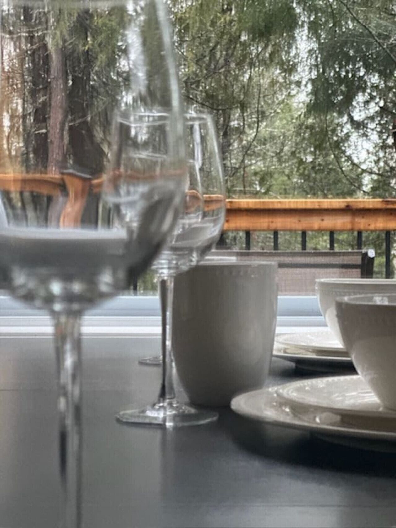 A table with a wine glass, a cup, and bowls, set against a backdrop of trees and a wooden railing.