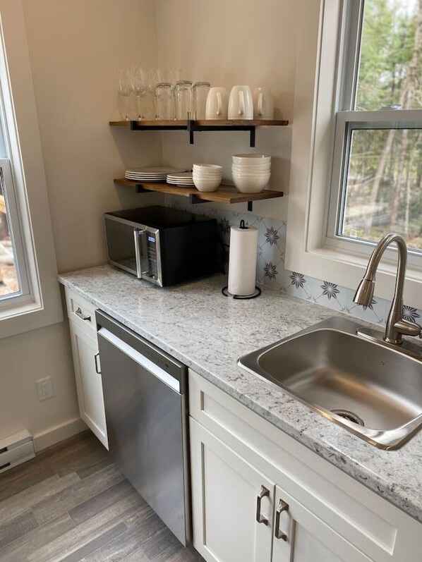 A kitchen with a stainless steel sink, a microwave, a dish rack, and a window with a view of trees. - Compass Cove Cottage with Private Hot tub (Waweig)