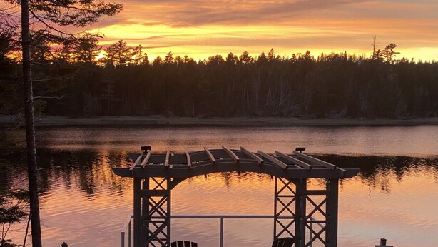 Ein hölzerner Pavillon mit zwei Stühlen und einem Tisch am Ufer eines stillen Sees bei Sonnenuntergang.
