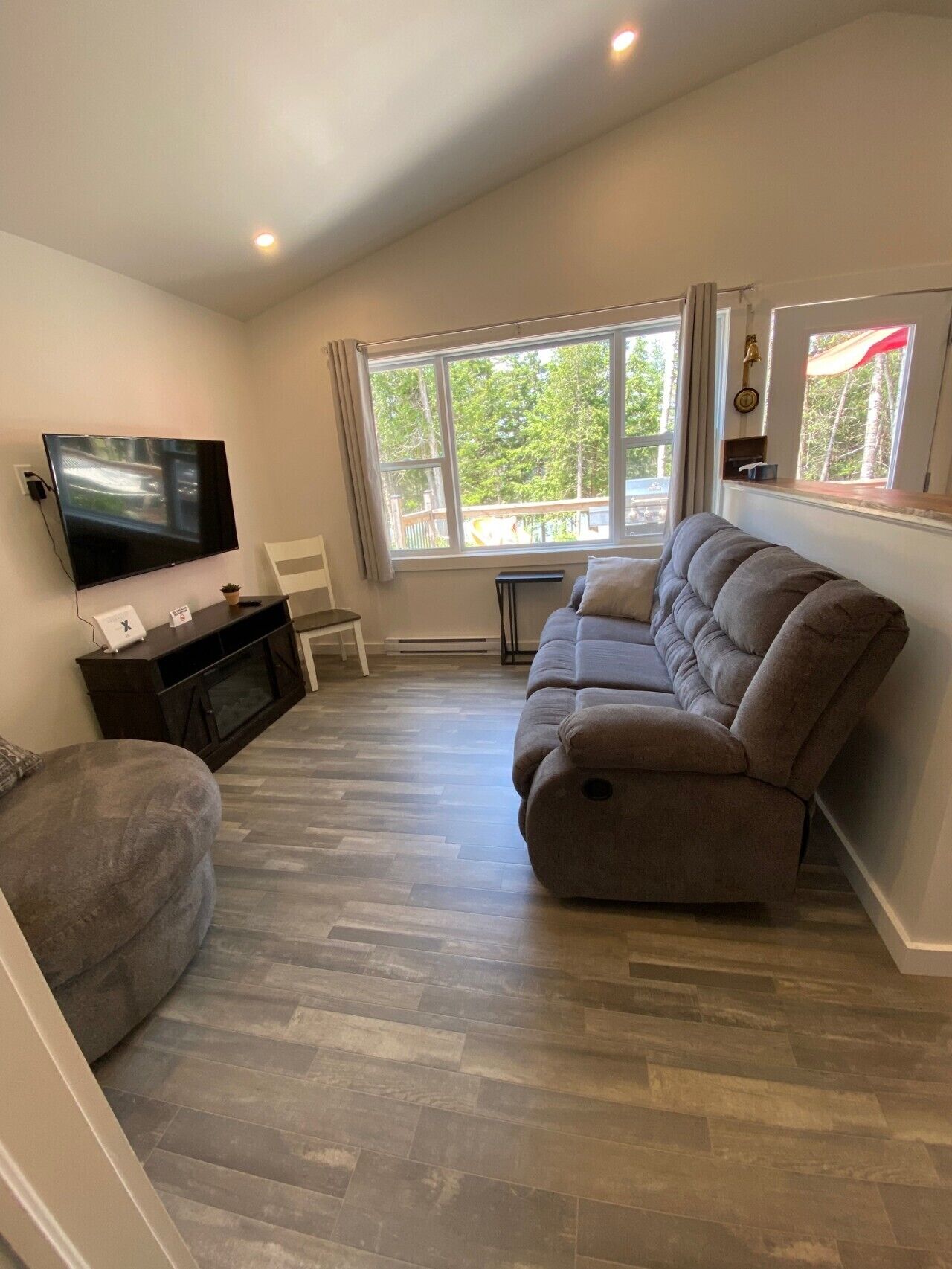A living room with a flat-screen TV, a sofa, a chair, and a window with a view of trees.