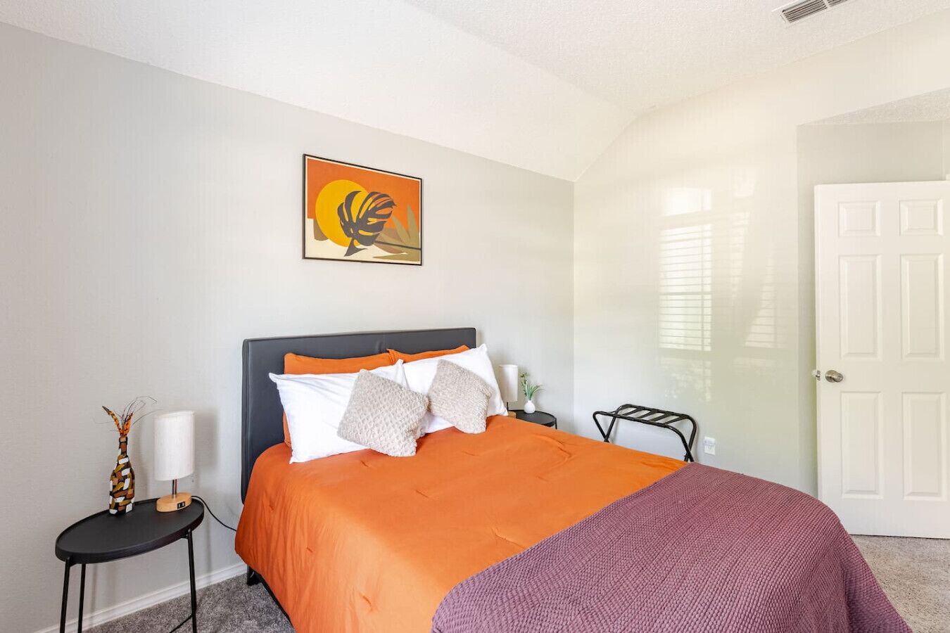 A bedroom with a bed, a nightstand, a lamp, a door, and a wall-mounted artwork.