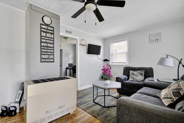 En moderne stue med ventilator, fladskærms-tv, en grå sofa og et lille bord med en vase med blomster.