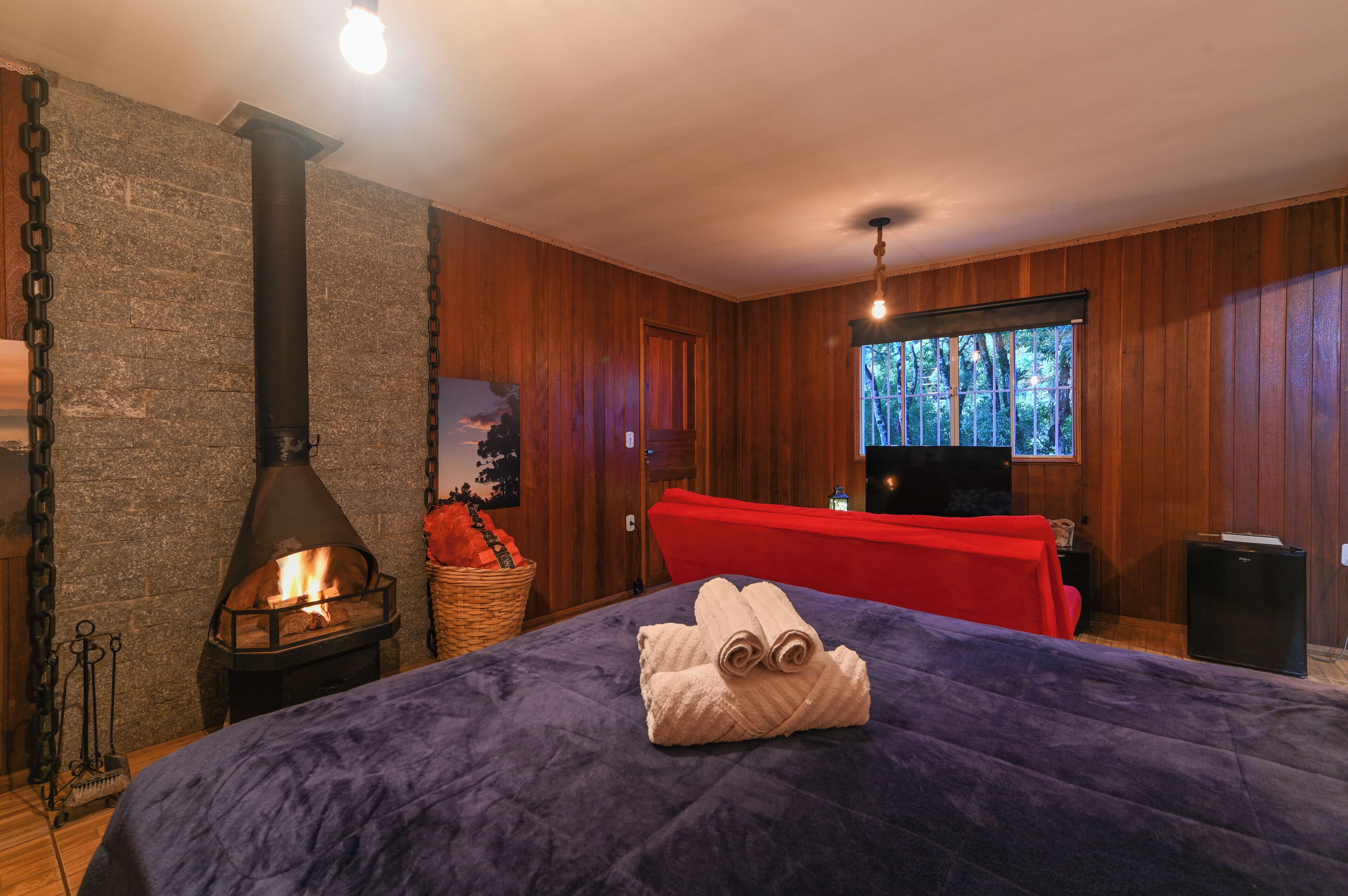 Um quarto acolhedor com parede de pedra, lareira a lenha, cama com manta roxa e uma cadeira vermelha.