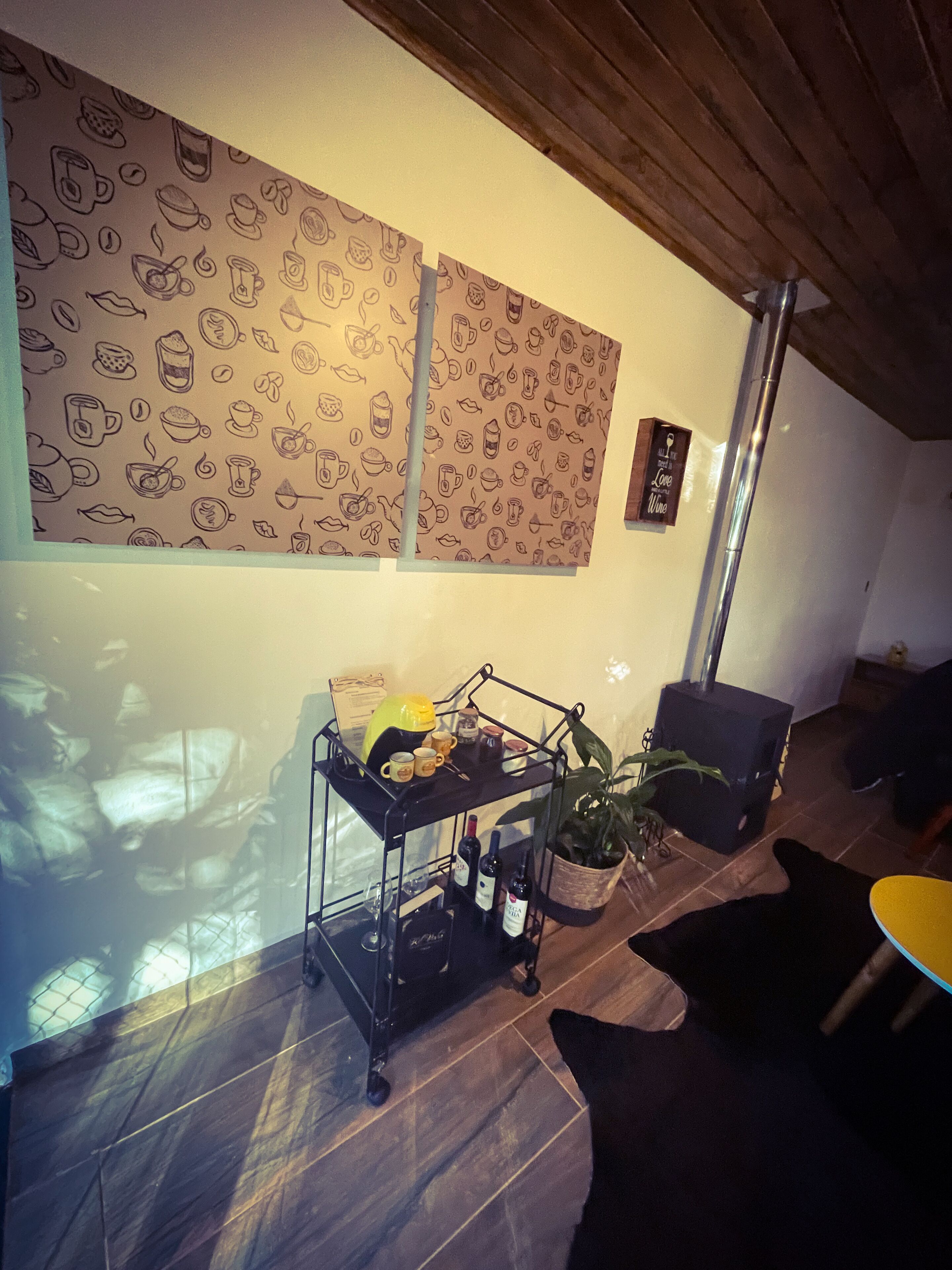 A cozy room with a wooden ceiling, a wall with a coffee-themed pattern, a metal cart with bottles, a potted plant, and a small table.