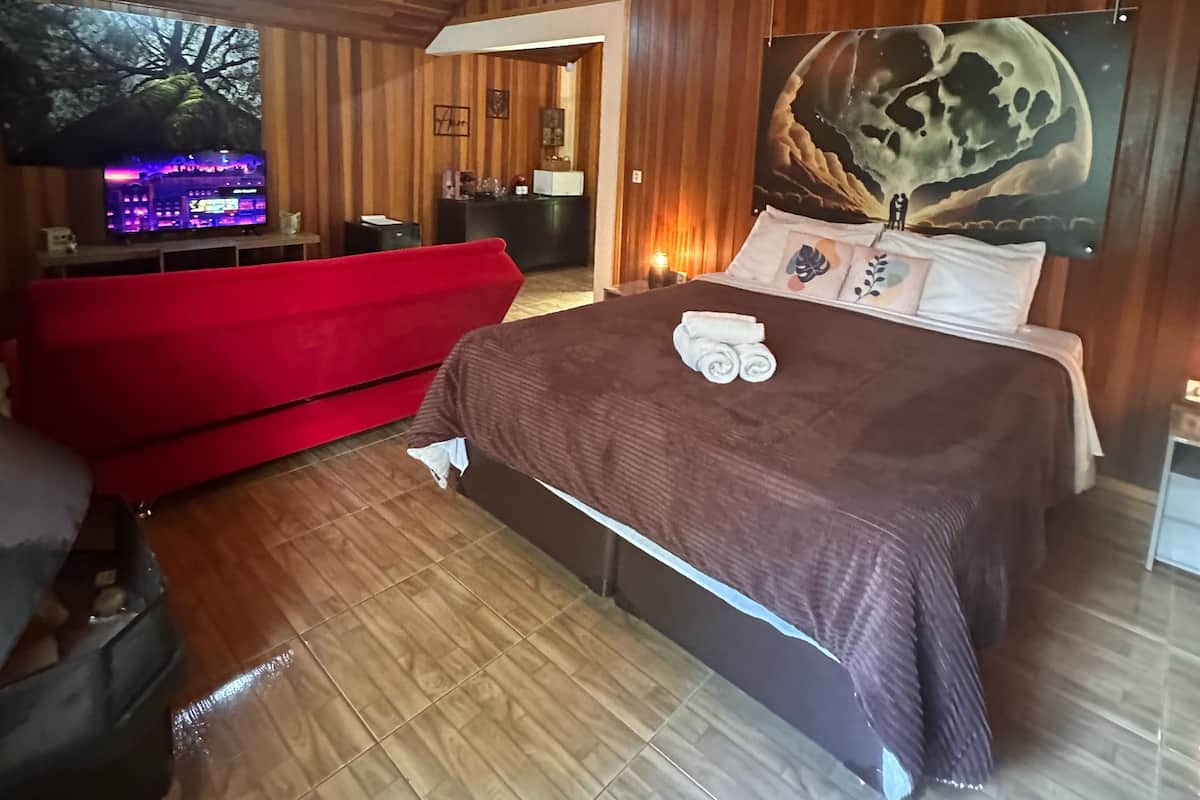 A bedroom with a wooden ceiling, a large bed with a brown blanket, a red sofa, and a painting of a moon.