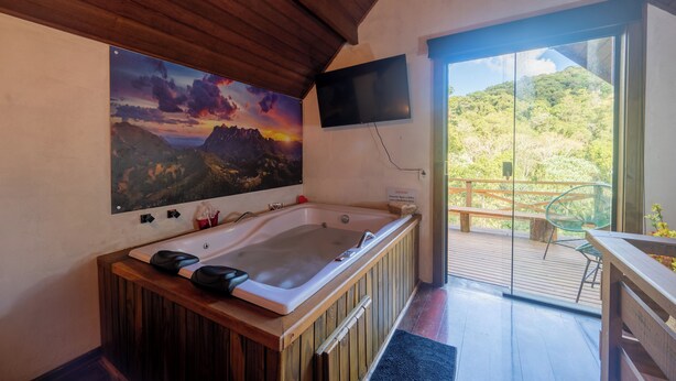 Un baño con una bañera de madera, un cuadro grande de un atardecer, un televisor montado en la pared y un balcón con vista a la vegetación.