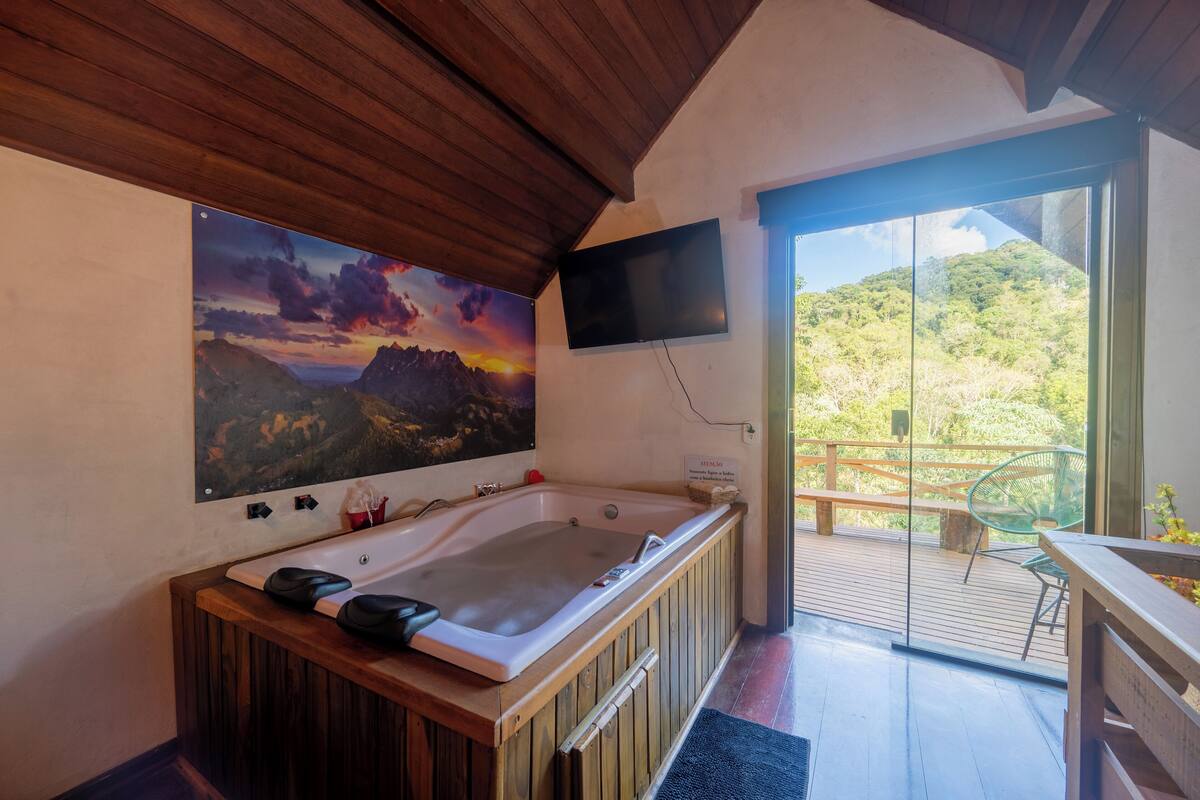 A bathroom with a wooden bathtub, a large painting of a sunset, a television mounted on the wall, and a balcony with a view of greenery.