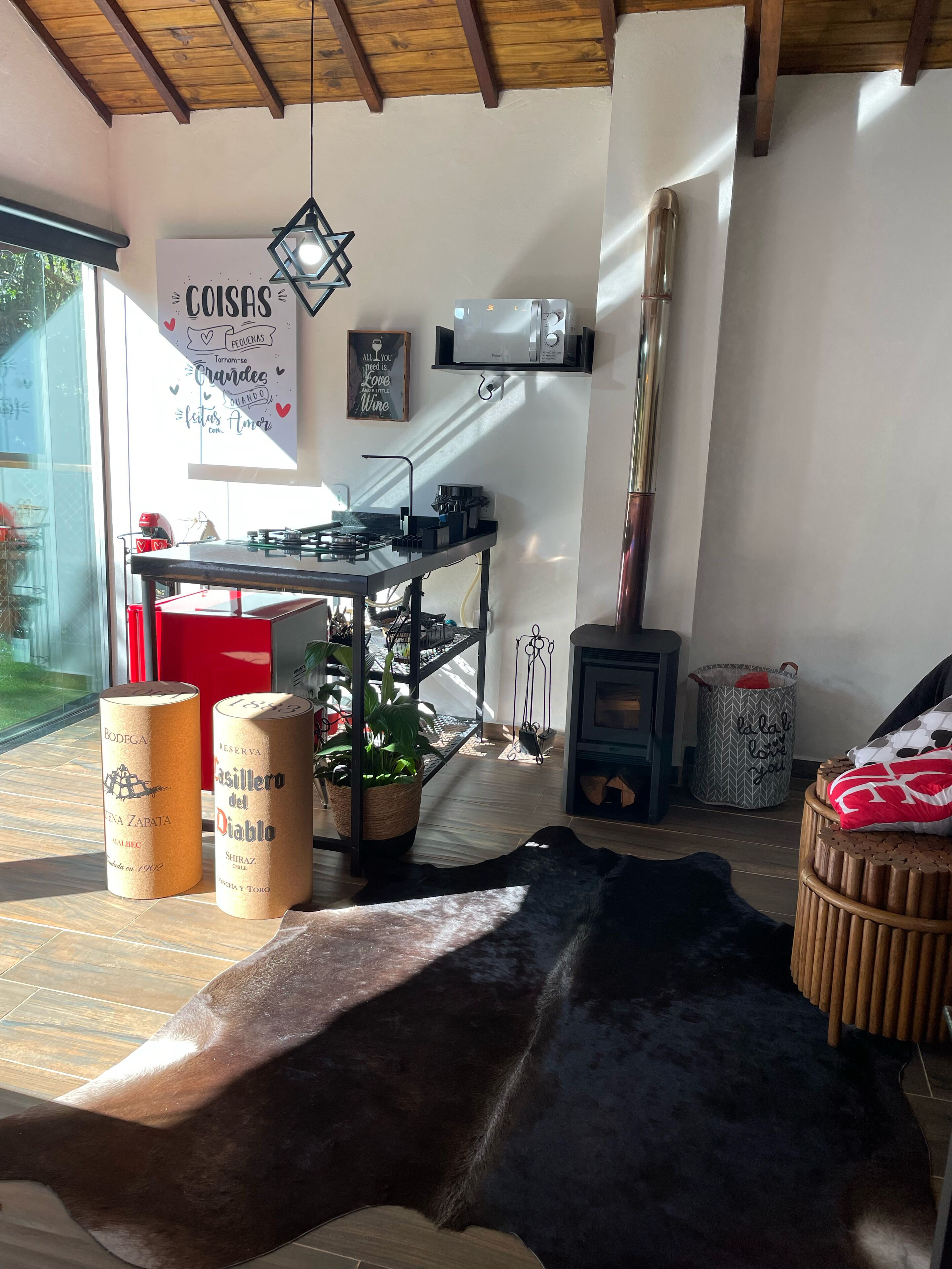 A cozy kitchen with a wooden ceiling, a gas stove, a microwave, and a hanging light fixture. There are two wine bottles labeled 'Bodega' and 'Casillero del Diablo' on the floor.