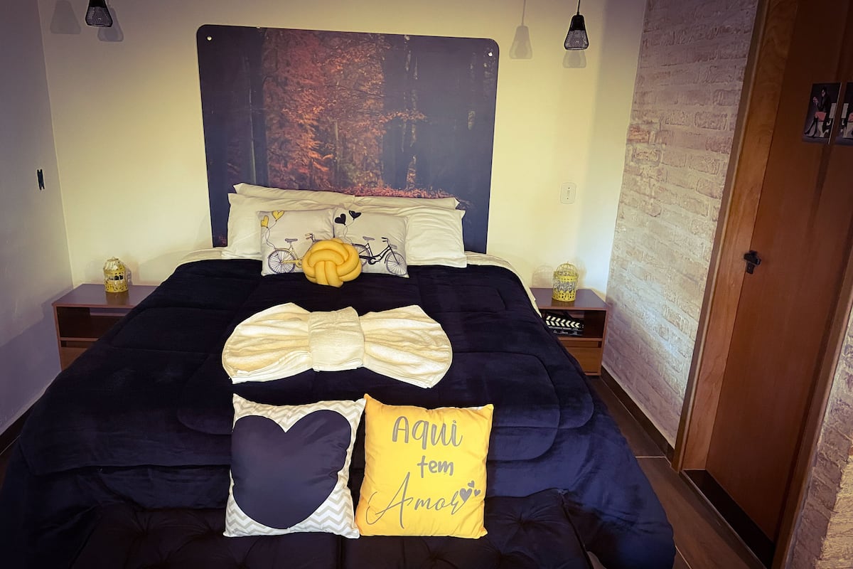 A neatly made bed with a large decorative bow pillow, a heart-shaped pillow, and a yellow pillow with text. There are two bedside tables with lamps and decorative items.