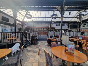 Un restaurante con techo alto, mesas y sillas de madera. Se observa una zona de bar al fondo.