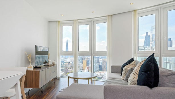 Una sala de estar moderna con sofá, una mesa de comedor y televisor. Amplias ventanas ofrecen vista a la ciudad.