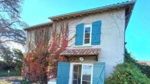 Una casa de dos pisos con contraventanas azules, techo de tejas y un pequeño patio con una mesa de vidrio.
