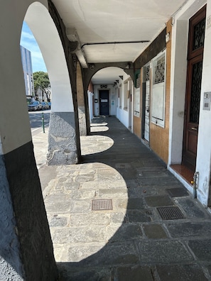 Un couloir au sol dallé, une porte au fond, et un bùtiment à la façade blanche avec une porte en bois sur le cÎté droit.